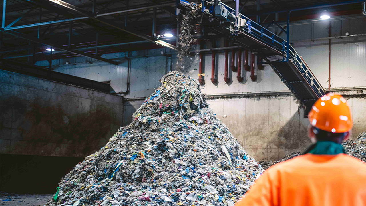 Rear view of young male worker in helmet, pollution mask, and reflective clothing observing waste falling from conveyor belt onto pile at facility.