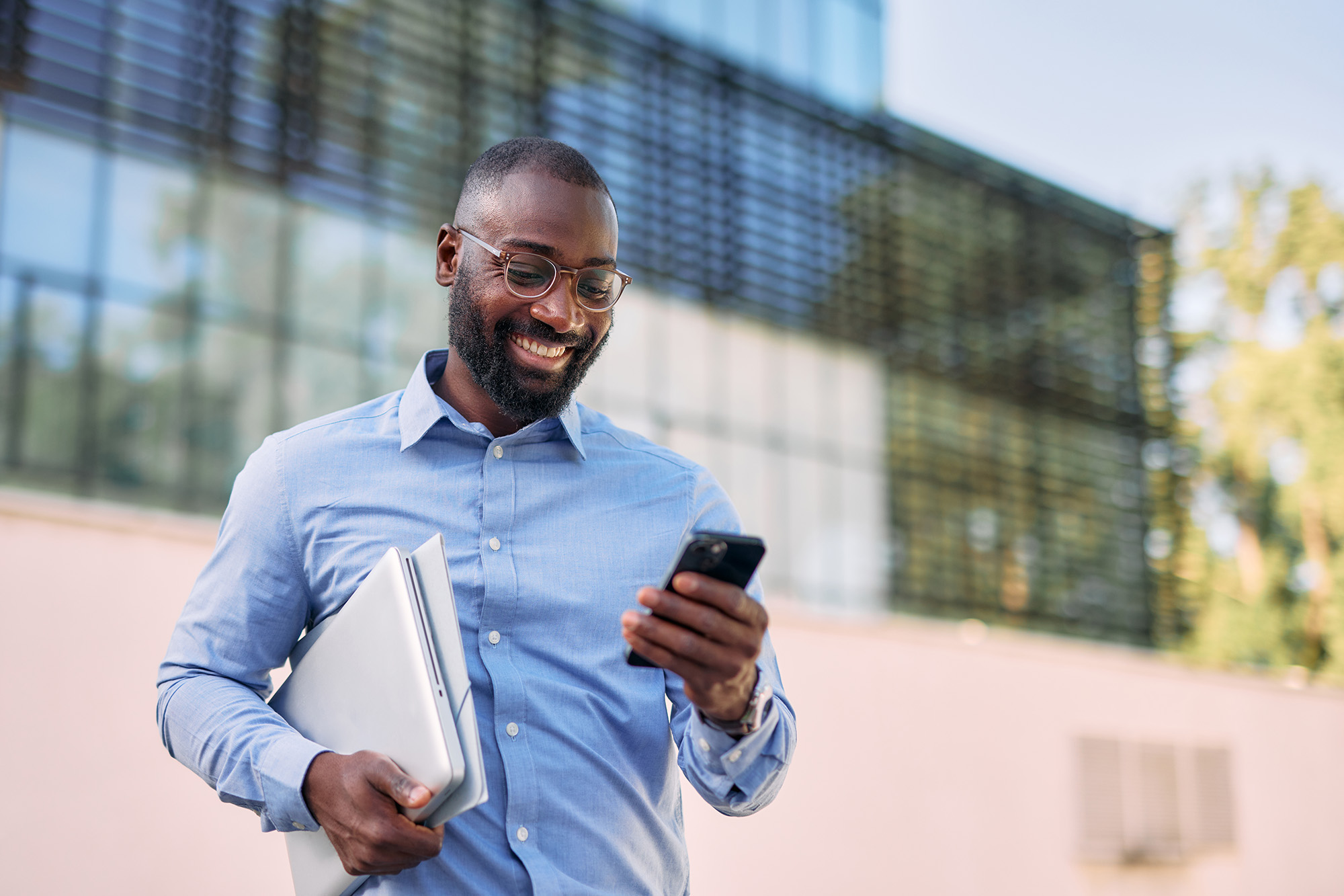 Smiling businessman using smartphone and carrying laptop in front of office building